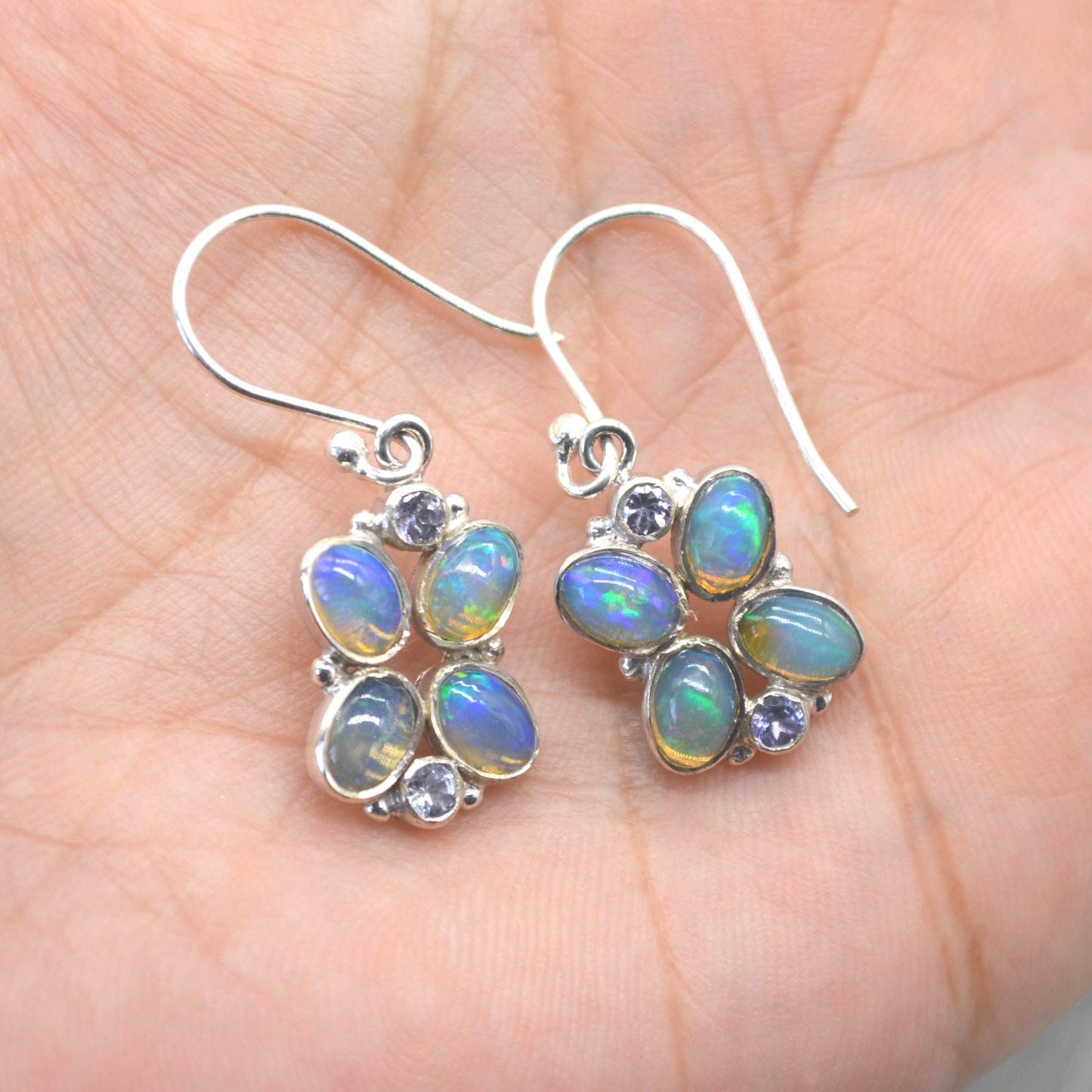 May include: Silver drop earrings with oval opals displaying iridescent blue and green colours. Each earring features a cluster of opals and small purple gemstones, forming a floral pattern. The earrings have a hook fastening.