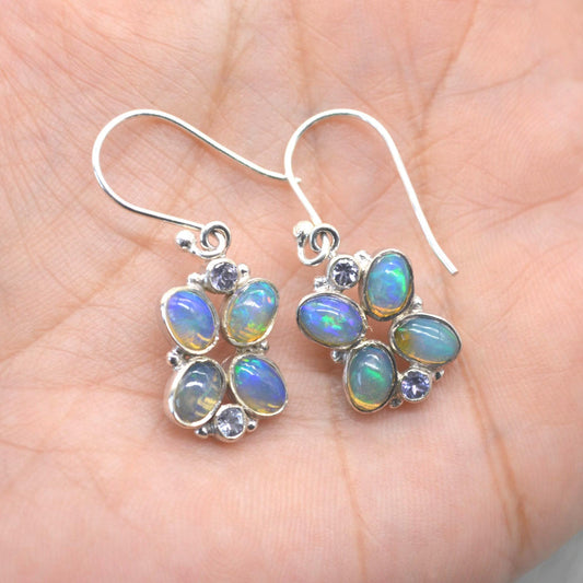 May include: Silver drop earrings with oval opals displaying iridescent blue and green colours. Each earring features a cluster of opals and small purple gemstones, forming a floral pattern. The earrings have a hook fastening.