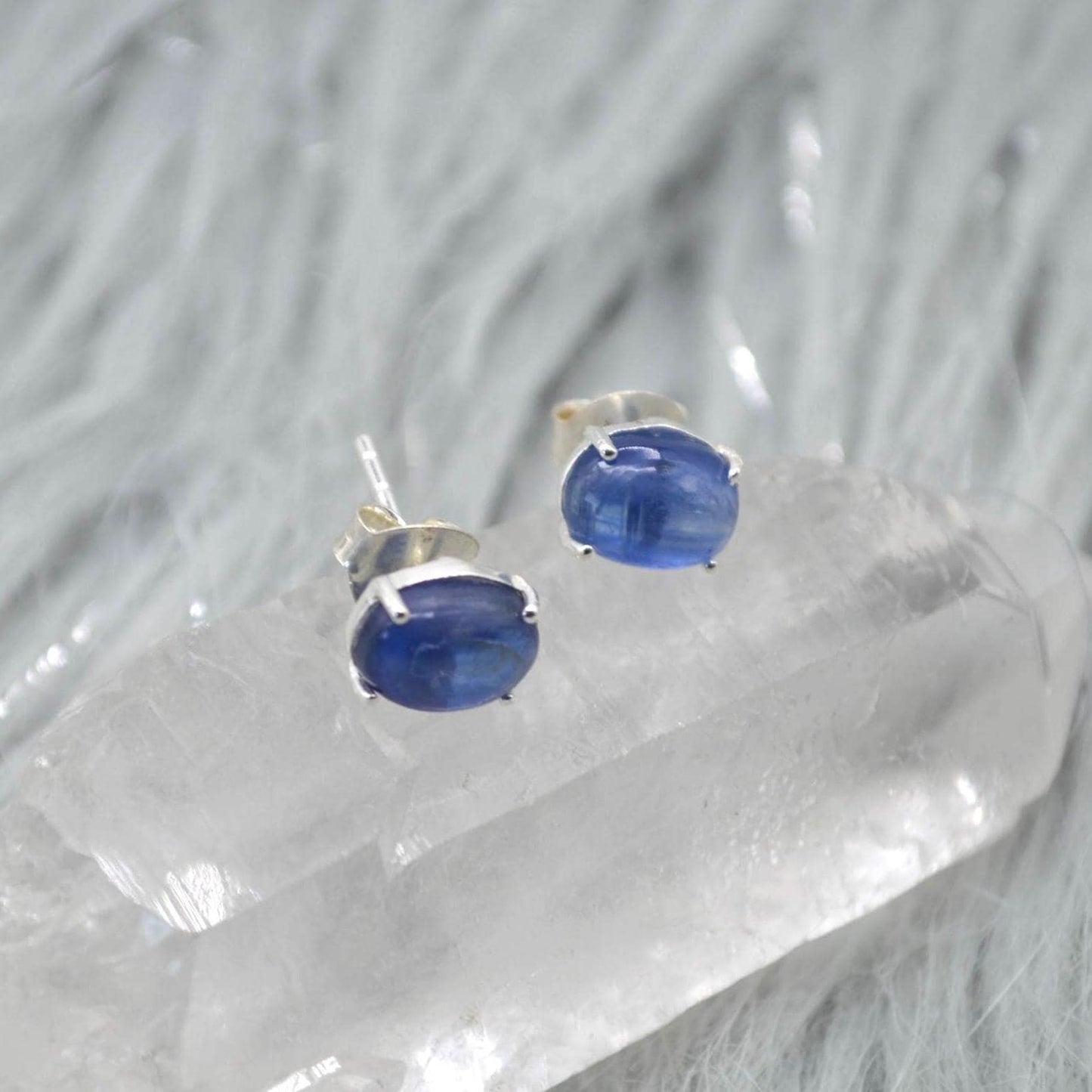 May include: A pair of oval blue kyanite stud earrings. Each earring has a polished, vibrant blue gemstone set in silver-coloured metal. The earrings are displayed on a clear, crystalline surface.