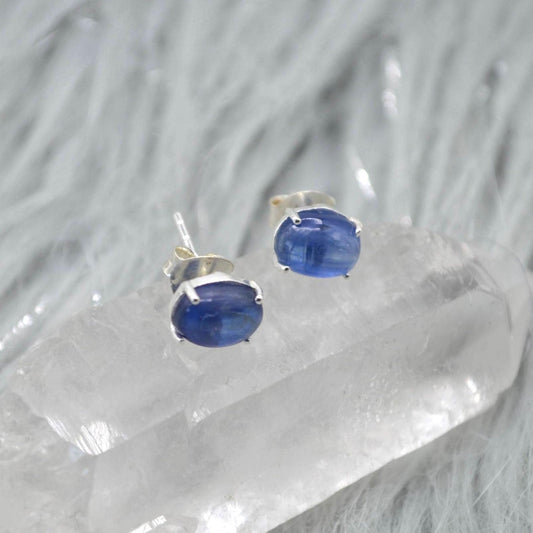 May include: A pair of oval blue kyanite stud earrings. Each earring has a polished, vibrant blue gemstone set in silver-coloured metal. The earrings are displayed on a clear, crystalline surface.